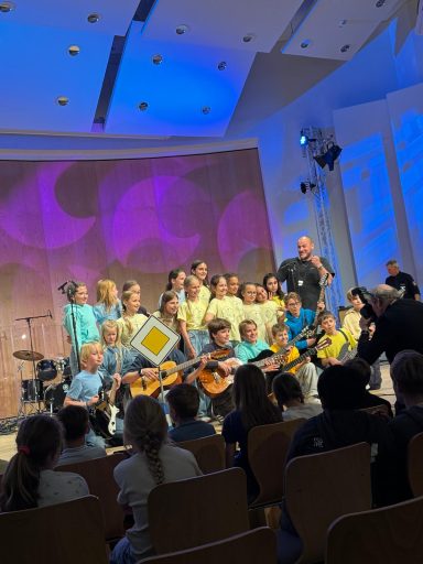 Kinderchor tritt auf, mit Instrumenten, vor buntem Hintergrund. Publikum blickt zu.