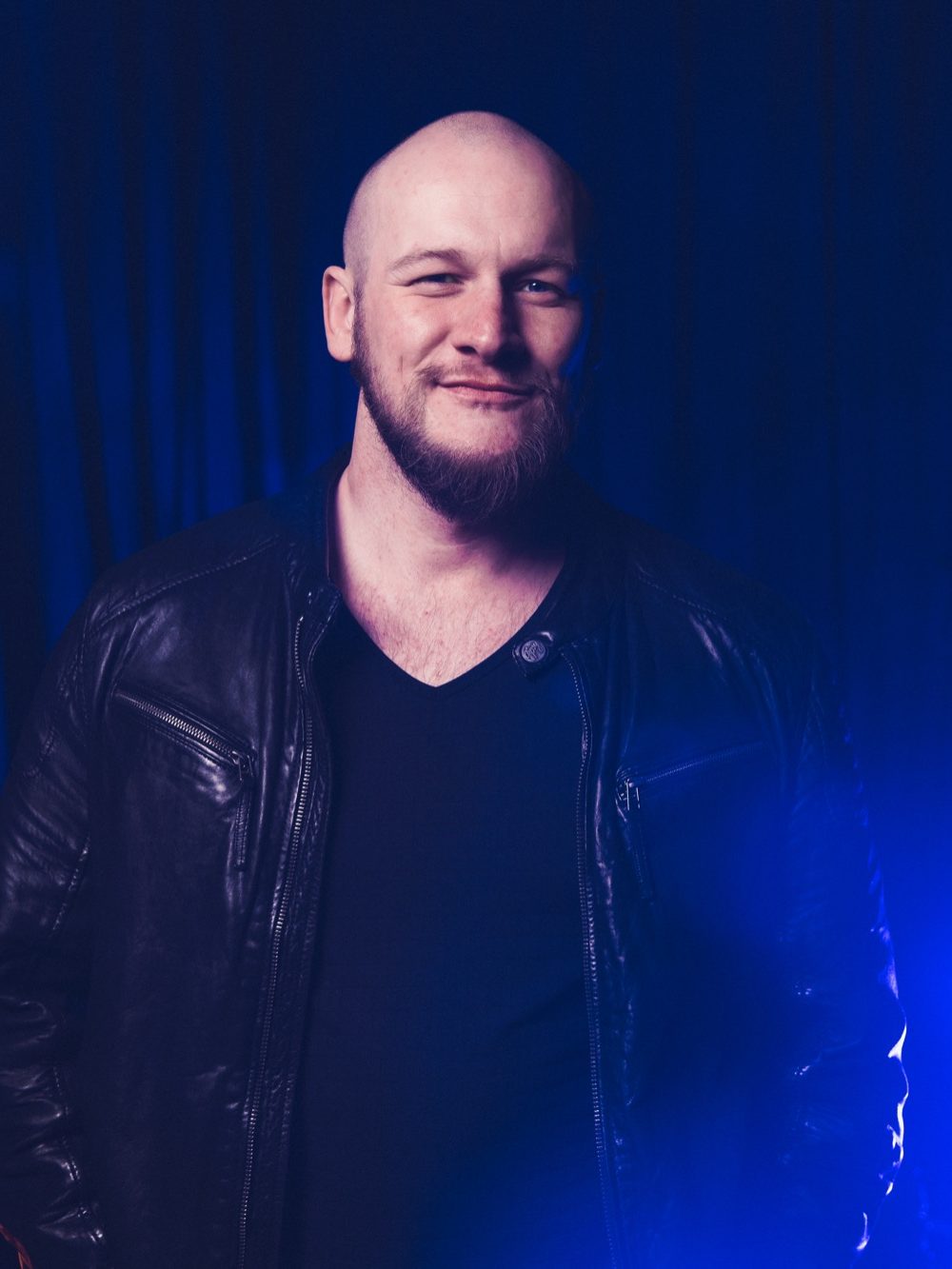 Mann mit Glatze und Bart, trägt eine schwarze Lederjacke, lächelt vor dunklem Hintergrund.